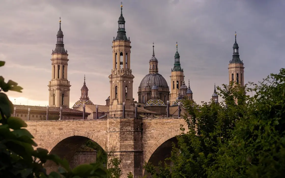 El perfil inconfundible de la Catedral-Bas&iacute;lica del Pilar, con c&uacute;pulas y torres elev&aacute;ndose hacia el cielo junto al Ebro, ha sido durante siglos una imagen de enorme significaci&oacute;n para los zaragozanos. El templo alberga una peque&ntilde;a columna de jaspe, el Pilar, que seg&uacute;n la tradici&oacute;n fue colocado por la Virgen Mar&iacute;a. De &eacute;l toma su nombre, y con &eacute;l nace un v&iacute;nculo entre el pueblo y su patrona que dura siglos.
La sensaci&oacute;n de grandeza y solemnidad nos acompa&ntilde;an desde que nos acercamos, y es que las dimensiones de la bas&iacute;lica impresionan. Dentro, los techos altos, con c&uacute;pulas pintadas por el mism&iacute;simo Goya, nos dejan con la boca abierta. Por todo ello, el Pilar es m&aacute;s que un monumento: es un lugar donde la fe y el arte se encuentran.