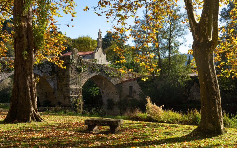 Un puente romano rodeado de vegetación y con el campanario de una iglesia de fondo
